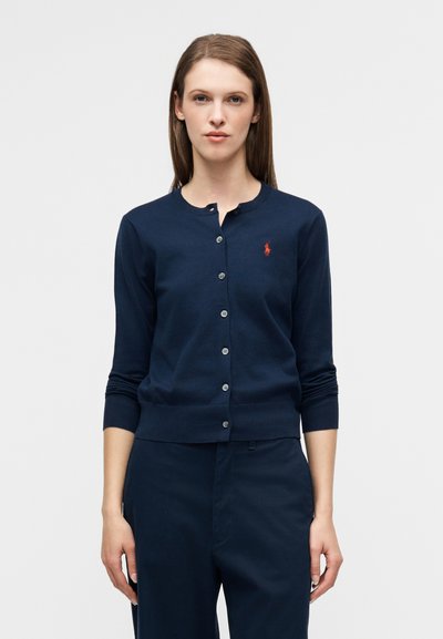 Καρδιάν navy μπλε σε ελαφρύ ύφασμα, με στρογγυλή λαιμόκοψη, κλείσιμο με κουμπιά και μικρό κόκκινο λογότυπο στο στήθος. Μακριά μανίκια.