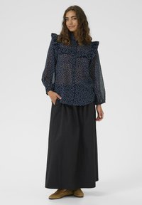 Nő tengerészkék áttetsző pöttyös blúzban, fodrokkal, fekete hosszú szoknyában és barna loaferekben, állva, az egyik kezével a zsebében.