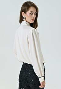 Blusa bianca fluida con maniche lunghe a palloncino, dettagli neri ai polsini, abbinata a pantaloni neri in paillettes, tessuto liscio, colletto elegante.