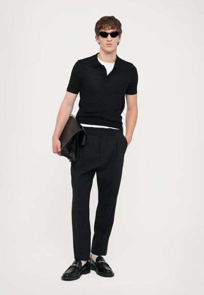 Crna pletiva polo majica, krojene crne hlače i sjajne crne cipele. Kožni clutch se drži u lijevoj ruci. Naočale za sunce upotpunjuju izgled.