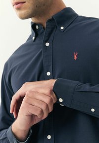 Camisa de botón marino con cuello puntiagudo, que presenta botones blancos y un pequeño logo de ciervo rojo en el pecho. Textura de algodón suave.