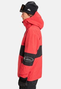 Rote und schwarze Skijacke mit Kapuze, mit einem Gittermuster, Logo-Detail auf dem Ärmel und einer strukturierten Oberfläche. Getragen mit einer Mütze und Skibrille.