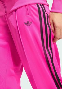 Fuchsia trainingsbroek met zwarte strepen en een geborduurd logo op de dij. Gemaakt van soepel, lichtgewicht materiaal en voorzien van zijzakken.
