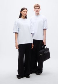 Φαρδιά μπλουζάκια σε ανοιχτό μπλε με ανάγλυφο λογότυπο, συνδυασμένα με μαύρο παντελόνι φαρδύ στο πόδι. Το μοντέλο κρατά μια μαύρη τσάντα με φερμουάρ.