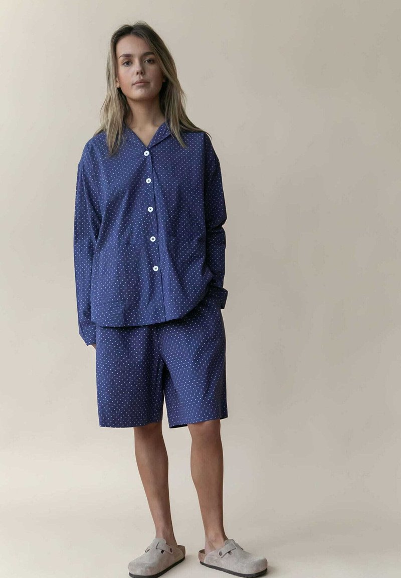 Navy-blaues Polka-Dot-Pyjama-Set mit einem langärmeligen Button-Up-Hemd und knielangen Shorts, kombiniert mit grauen Slippers.
