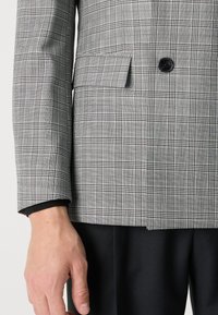 Grå rutigt blazer med notch-lapel, enkel knappstängning och framficka, kompletterad med en svart undertröja vid ärmslutet.