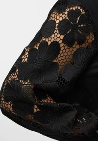 Blusa nera con maniche in pizzo con ricami floreali su uno sfondo di tessuto solido, che mette in mostra intricate fantasie e trame.