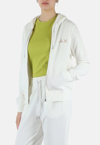 Felpa bianca con zip e cappuccio, dotata di laccetti con perline e un piccolo logo beige. Indossata con una maglietta verde aderente e pantaloni coordinati.