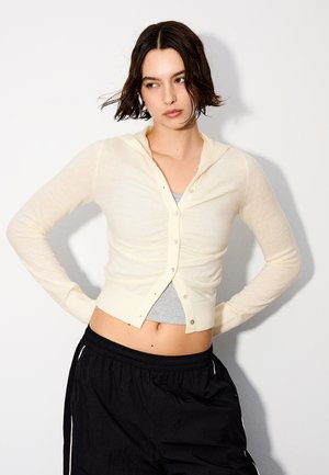 Frau mit kurzen dunklen Haaren trägt eine cremefarbene, geknöpfte Strickjacke über einem grauen Crop-Top und schwarzen Hosen, steht mit Händen in die Hüften gestützt vor weißem Hintergrund.