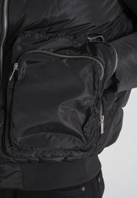 Giacca a vento nera con una texture lucida e liscia, dotata di una grande tasca con zip sul petto e polsini a coste sull'orlo.