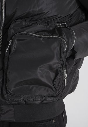 Černá péřová bunda s lesklou, hladkou texturou, která má velkou kapsu na zip na hrudi a pružné manžety na spodním lemu.