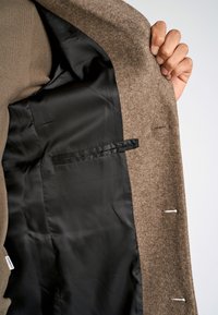 Intérieur en laine marron avec doublure en satin noir, featuring a chest pocket et des accents de couture visibles. Main tenant le bord du manteau.
