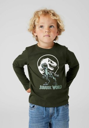 Blond child with curly hair wearing a dark green "Jurassic World" shirt with dinosaur graphics and blue jeans, hands behind back.