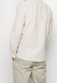 Camisa de manga larga de color beige claro hecha de lino, con un espalda plisada, puños abotonados y un dobladillo ligeramente curvado. Combinada con pantalones claros.