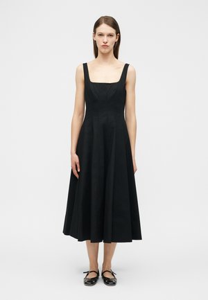 Kobieta ubrana w czarną, bezrękawową sukienkę midi z kwadratowym dekoltem oraz czarne baleriny, stojąca na tle jednolitego jasnego tła.