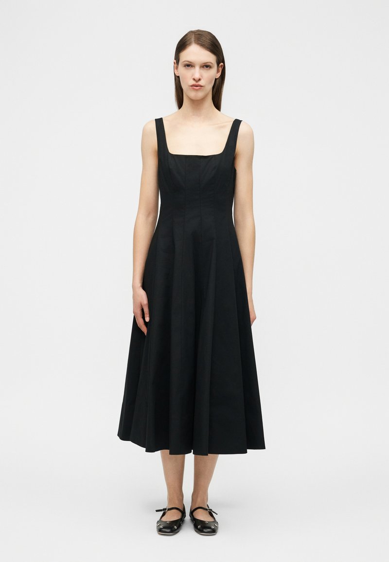 Γυναίκα που φοράει μαύρο μίντι φόρεμα χωρίς μανίκια με τετράγωνο ντεκολτέ και μαύρες μπαλαρίνες, στέκεται μπροστά σε απλό ανοιχτό φόντο.