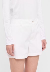 Shorts en coton blanc avec une taille ajustée, des poches latérales, un ourlet replié et une fermeture à bouton unique sur le devant. Associés à un haut blanc à manches longues.