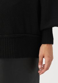 Pull en tricot noir avec des poignets et un ourlet côtelés. Texture douce et coupe décontractée. Main reposant à côté d'un matériau noir lisse sous le pull.