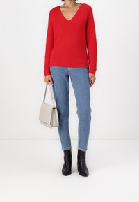 Maglione rosso a collo a v lavorato a maglia con maniche lunghe, abbinato a jeans skinny blu chiaro e stivaletti neri alla caviglia. Tiene in mano una borsa grigia chiaro.