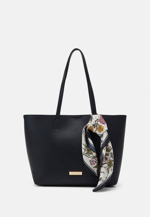 Black leather tote bag with two shoulder straps, featuring a floral silk scarf tied on the handle and a metallic logo accent on the front.