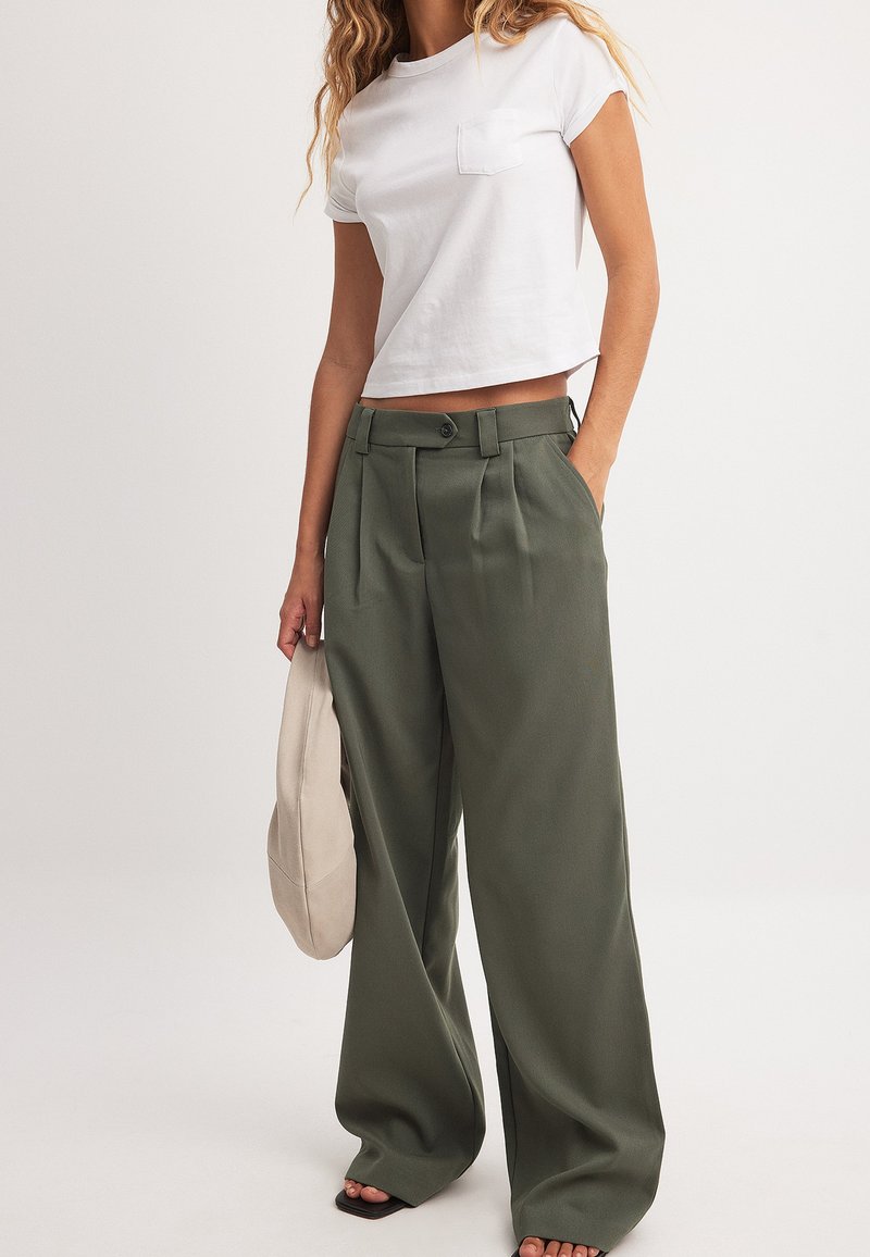 Bela skrajšana majica s kratkimi rokavi in žepom na prsih, v kombinaciji s širokimi, visokim pasom olivno zelenimi hlačami, ki imajo gube in sprednji gumb.