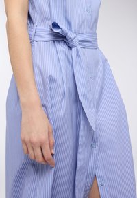 Abito camicia a righe blu con cintura a bretella, chiusura con bottoni e maniche corte. Il tessuto è liscio e con una leggera lucentezza.