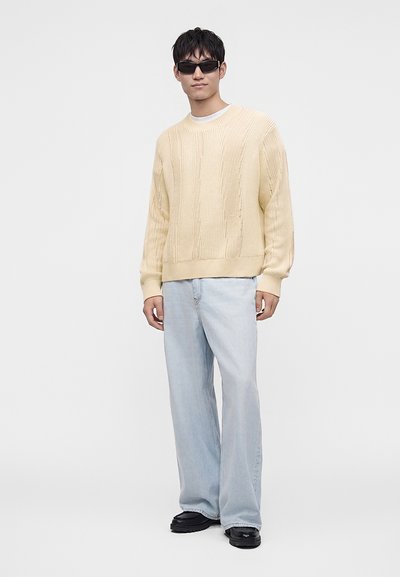 Jeune homme portant des lunettes de soleil noires, un pull en maille crème, un jean bleu clair large et des chaussures noires, debout devant un fond blanc.