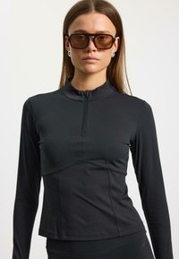 Žena oblečená v čiernom topánok s dlhým rukávom a polovičným zipsom, s hnedými oversized slnečnými okuliarmi a striebornými okrúhlymi náušnicami na jednoduchej pozadí.
