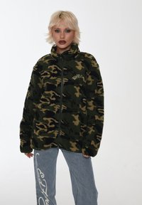 Giacca in pile camouflage verde e beige con colletto alto e zip completa, con testo ricamato sul lato sinistro del petto. Abbinata a jeans blu.