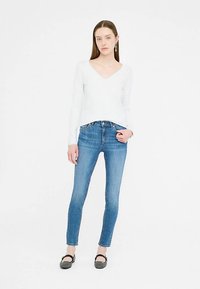 Pulover alb cu mâneci lungi și decolteu în V, asortat cu blugi skinny albaștri, cu talie medie, completat de pantofi negri, pozând pe un fundal simplu.