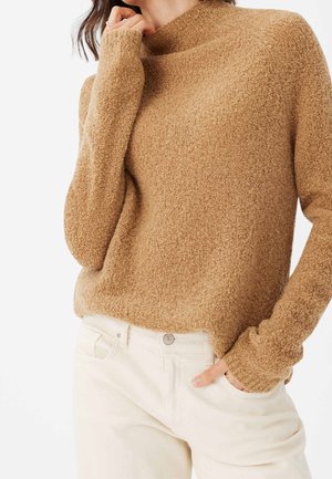Brauner, strukturierter Pullover mit hohem Kragen und langen Ärmeln, kombiniert mit hellfarbigen, schmal geschnittenen Hosen. Weicher, warmer Stoff.