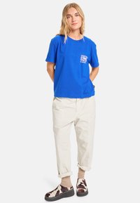 Blaues Baumwoll-T-Shirt mit einem kleinen grafischen Taschendesign, kombiniert mit hellbeigen Hosen und gemusterten Schuhen. Hochgekrempelte Ärmel und lässige Passform.