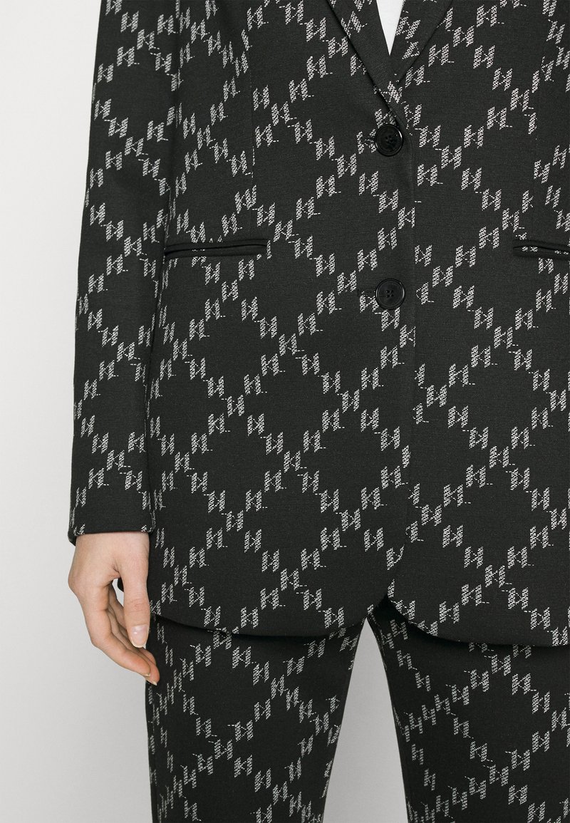 Abito nero su misura con motivo a zigzag bianco, due bottoni, tasche a pattina e pantaloni coordinati indossato da una persona con la mano rilassata al lato.