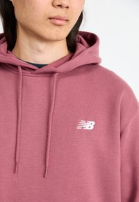 Rúžová mikina z jemnej látky, s klokaním vreckom, šnúrkami na stiahnutie a bielym logom New Balance na hrudi.