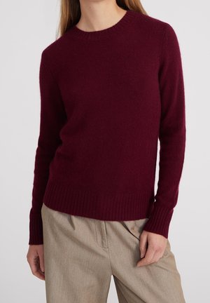 Kvinde iført en tætsiddende, langærmet, rundhalset bordeauxrød sweater med beige, højtaljede bukser, stående mod en ensfarvet lys baggrund.