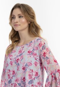 Blusa floreale azzurra chiaro con motivi di rose rosa e viola, collo rotondo e maniche lunghe a campana con una texture morbida e fluida.