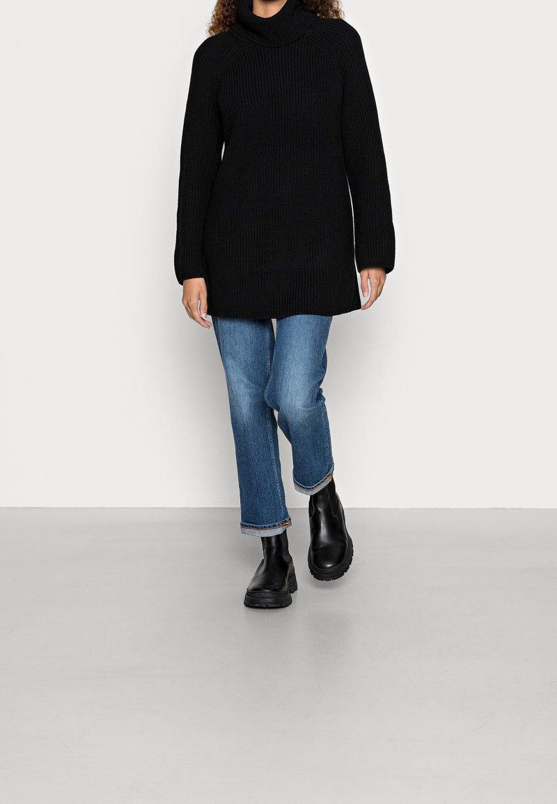 Personne portant un pull noir à col roulé à manches longues, un jean bleu avec revers et des bottines noires, marchant vers l'avant sur un fond clair uni.