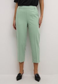 Pantaloni su misura verde menta con un design cropped, caratterizzati da pieghe frontali e una trama liscia. Abbinati a scarpe nere con punta affusolata.