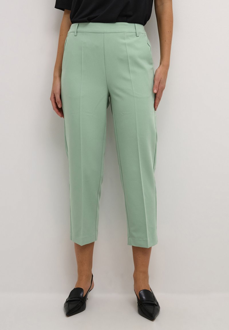 Pantaloni su misura verde menta con un design cropped, caratterizzati da pieghe frontali e una trama liscia. Abbinati a scarpe nere con punta affusolata.