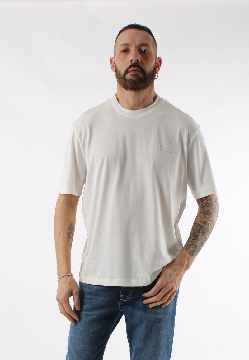 T-shirt in cotone color crema con maniche corte e scollatura rotonda, decorato con un piccolo logo stampato sul lato sinistro. Abbinato a jeans blu.