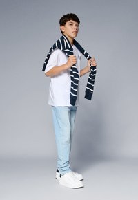 T-shirt bianca a maniche corte, jeans azzurri chiari e sneakers bianche. Un maglione a strisce blu navy è casualmente appoggiato sulle spalle.
