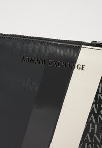 Crna ziperska torbica od teksturiranog materijala, s natpisom "ARMANI EXCHANGE" u metalik slovima. Geometrijski crno-bijeli uzorak s jedne strane.