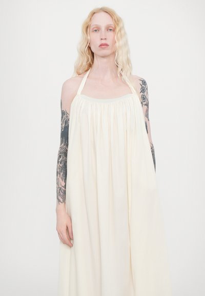 Ljuskräm halterneck-kjol med veckad tyg vid bysten, flödande silhuett och en mjuk textur. Accentuerad av synliga tatueringar på armarna.