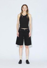 Top sin mangas negro y pantalones cortos anchos negros de mezclilla, con bolsillos frontales y herrajes de botón marrón claro. Combinado con calcetines crema y zapatos negros.