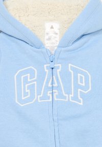 Svetlomodrá mikina na zips vyrobená z mäkkého materiálu, s krémovou fleecovou podšívkou a veľkými bielymi nápismi "GAP" na prednej strane.