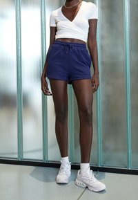 Haut court blanc à effet drapé, associé à un short bleu marine à cordon de serrage, complété par des baskets blanches de sport et des chaussettes chevilles.