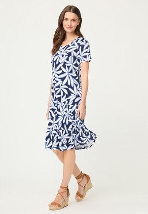 Kobieta w granatowo-białej sukience w kwiaty z krótkim rękawem i brązowych sandałach na koturnie, stoi z jedną lekko uniesioną nogą.