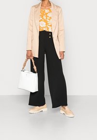 Béžový blazer přes květinový žlutý top, černé široké kalhoty se zlatými knoflíky, doplněné bílou kabelkou a béžovými platformovými botami.