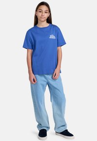 Camiseta de algodón azul con mangas cortas, que presenta un logotipo en el pecho. Combinada con pantalones vaqueros holgados de color azul claro y zapatillas oscuras.