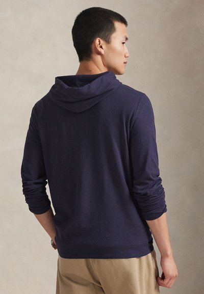 Mees, kes seisab küljepealt, kannab tumesinist pika varrukaga kapuutsi ja beeže pükse ühtlasel heledal taustal.